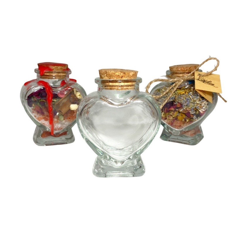 May include: Three heart-shaped glass jars with cork stoppers. Two of the jars are filled with dried flowers and herbs, while the third jar is empty. The jars are perfect for storing small items or creating unique gifts.