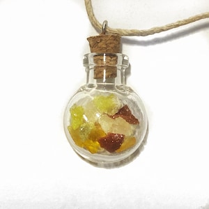 Può includere: Una piccola bottiglia di vetro con un tappo di sughero riempita di pezzi di vetro giallo, arancione e marrone. La bottiglia è attaccata a una collana di corda marrone.