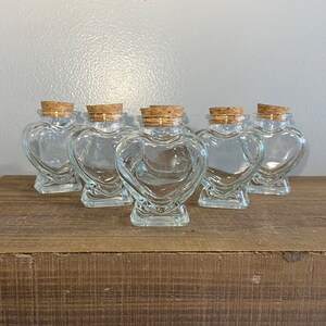 May include: Six clear glass heart-shaped jars with cork lids. The jars are arranged in a row on a wooden surface.
