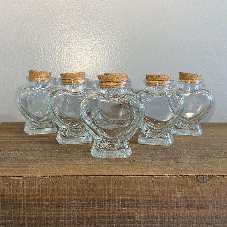 May include: Six clear glass heart-shaped jars with cork lids. The jars are arranged in a row on a wooden surface.