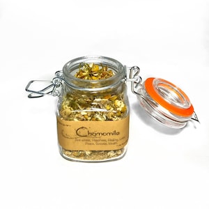 Puede incluir: Un frasco de vidrio con tapa de metal lleno de flores de manzanilla secas. El frasco tiene una etiqueta de papel marrón que dice "Chamomile" y enumera beneficios como "Anti-estrés, Felicidad, Sanación, Amor, Paz, Éxito, Riqueza".