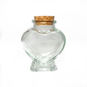 May include: A clear glass heart-shaped bottle with a cork stopper. The bottle is empty and has a smooth, rounded surface.