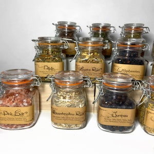 Puede incluir: Una colecci&oacute;n de diez frascos de vidrio con etiquetas de papel marr&oacute;n y tapas naranjas. Los frascos contienen varias hierbas y especias, incluyendo ra&iacute;z de regaliz, ra&iacute;z de malvavisco, enebro, sal rosa y bayas de sa&uacute;co. Los frascos est&aacute;n dispuestos sobre una superficie de madera.