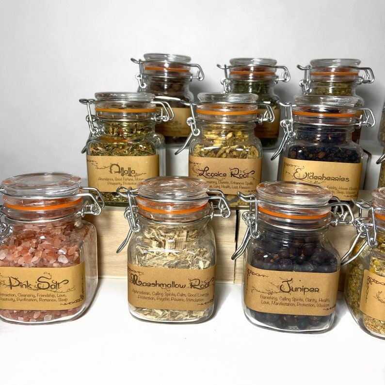 Puede incluir: Una colecci&oacute;n de diez frascos de vidrio con etiquetas de papel marr&oacute;n y tapas naranjas. Los frascos contienen varias hierbas y especias, incluyendo ra&iacute;z de regaliz, ra&iacute;z de malvavisco, enebro, sal rosa y bayas de sa&uacute;co. Los frascos est&aacute;n dispuestos sobre una superficie de madera.