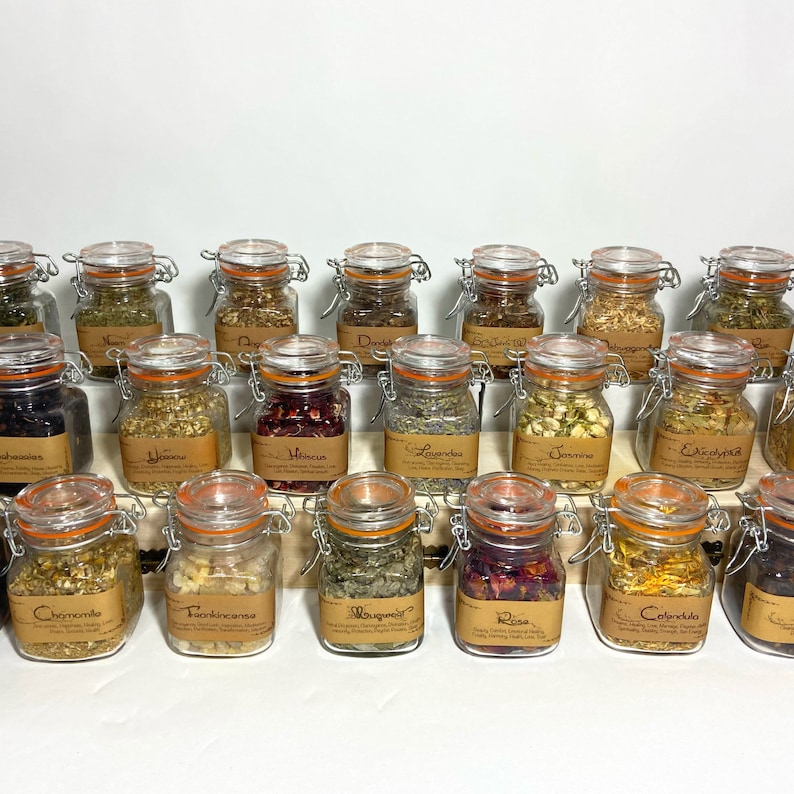 Puede incluir: Una colecci&oacute;n de 18 frascos de vidrio con diversas hierbas y flores secas. Los frascos est&aacute;n dispuestos en dos filas en un estante de madera. Cada frasco tiene una etiqueta con el nombre de la hierba o la flor. Las hierbas y las flores incluyen manzanilla, incienso, artemisa, rosa, cal&eacute;ndula, hibisco, lavanda, jazm&iacute;n, equin&aacute;cea, y m&aacute;s.