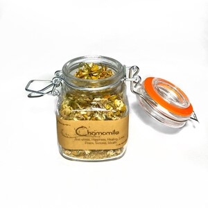 Puede incluir: Un frasco de vidrio con tapa de metal lleno de flores de manzanilla secas. El frasco tiene una etiqueta de papel marr&oacute;n que dice "Chamomile" y enumera beneficios como "Anti-estr&eacute;s, Felicidad, Sanaci&oacute;n, Amor, Paz, &Eacute;xito, Riqueza".