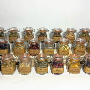 Puede incluir: Una colecci&oacute;n de 20 frascos de vidrio con etiquetas y tapas, llenos de hierbas y flores secas. Los frascos est&aacute;n dispuestos en filas sobre una superficie blanca. Las etiquetas incluyen nombres como "Manzanilla", "Hibisco", "Lavanda" y "Rosa".