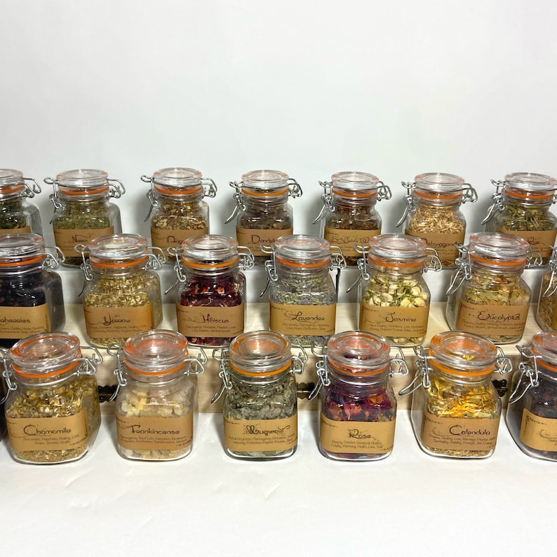 Puede incluir: Una colecci&oacute;n de 20 frascos de vidrio con etiquetas y tapas, llenos de hierbas y flores secas. Los frascos est&aacute;n dispuestos en filas sobre una superficie blanca. Las etiquetas incluyen nombres como "Manzanilla", "Hibisco", "Lavanda" y "Rosa".