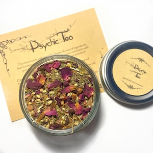 Op de afbeelding: Een glazen pot gevuld met een mengsel van gedroogde kruiden, specerijen en rozenblaadjes. De pot is gelabeld "Psychic Tea" en staat op bruin papier met tekst die luidt "This tea is made with...chosen all-natural ingredients that boost your connection with your practices!"