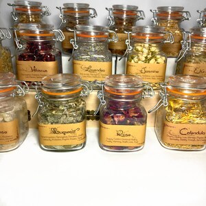 Puede incluir: Una colecci&oacute;n de peque&ntilde;os frascos de vidrio con etiquetas de papel marr&oacute;n y tapas naranjas. Los frascos contienen hierbas y flores secas. Las etiquetas enumeran los nombres de las hierbas y flores, incluyendo hibisco, lavanda, jazm&iacute;n, artemisa, rosa y cal&eacute;ndula.