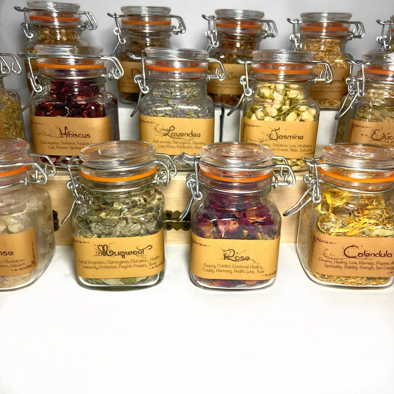 Puede incluir: Una colecci&oacute;n de peque&ntilde;os frascos de vidrio con etiquetas de papel marr&oacute;n y tapas naranjas. Los frascos contienen hierbas y flores secas. Las etiquetas enumeran los nombres de las hierbas y flores, incluyendo hibisco, lavanda, jazm&iacute;n, artemisa, rosa y cal&eacute;ndula.