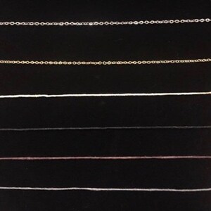 Op de afbeelding: Een close-up van zes verschillende soorten kettingen, waaronder zilver, goud, wit, zwart, bruin en wit. De kettingen zijn in rijen op een zwarte achtergrond gerangschikt.