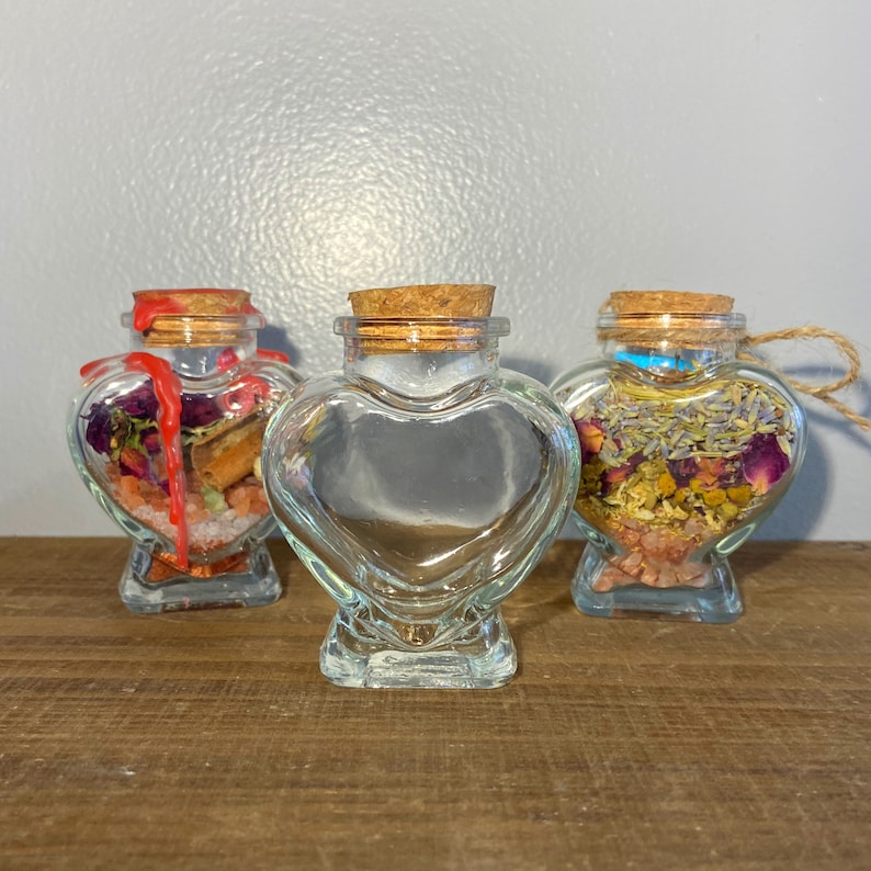May include: Three heart-shaped glass jars with cork stoppers. Each jar is filled with dried herbs and flowers. The jars are on a wooden surface.