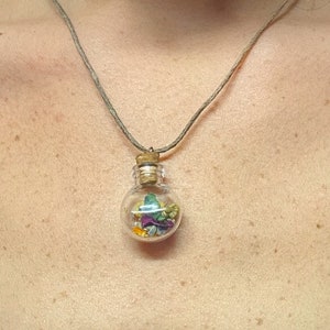 Op de afbeelding: Een delicate ketting met een klein glazen flesje gevuld met gedroogde bloemen. Het flesje hangt aan een bruin koord.