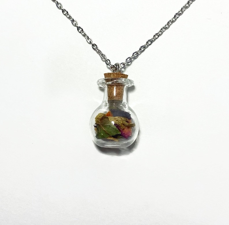 Op de afbeelding: Een zilveren ketting met een klein glazen flesje als hanger, gevuld met gedroogde bloemen en bladeren. De fles heeft een kurken stop.