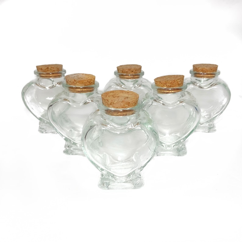 May include: Six clear glass heart-shaped bottles with cork stoppers. The bottles are arranged in a semi-circle on a white background.