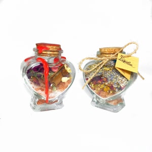 May include: Two heart-shaped glass jars filled with dried flowers, herbs, and salt. The jar on the left has a red wax seal. The jar on the right has a brown twine bow and a yellow tag that says "Spell Love".