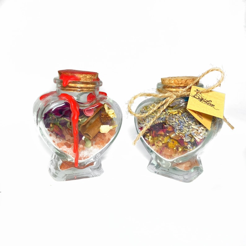May include: Two heart-shaped glass jars filled with dried flowers, herbs, and salt. The jar on the left has a red wax seal. The jar on the right has a brown twine bow and a yellow tag that says "Spell Love".