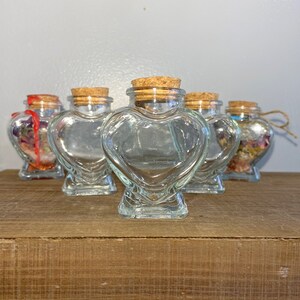 May include: Five clear glass heart-shaped bottles with cork stoppers. The bottles are arranged in a row on a wooden surface.
