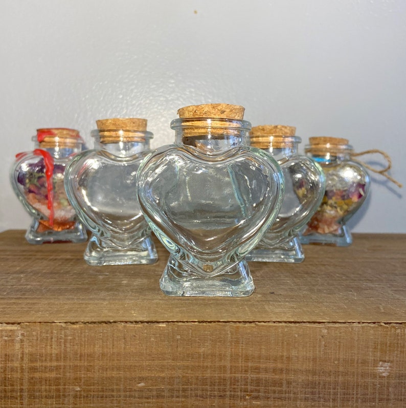 May include: Five clear glass heart-shaped bottles with cork stoppers. The bottles are arranged in a row on a wooden surface.