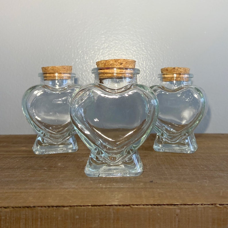 May include: Three clear glass heart-shaped jars with cork lids. The jars are empty and sitting on a wooden surface.