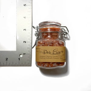 Puede incluir: Un peque&ntilde;o frasco de vidrio con tapa de cierre met&aacute;lico lleno de sal rosa del Himalaya. El frasco tiene una etiqueta de papel marr&oacute;n que dice "Pink Salt" y enumera los beneficios de la sal, incluyendo la atracci&oacute;n, la limpieza, la amistad, el amor, la positividad, la purificaci&oacute;n, el romance y el sue&ntilde;o.
