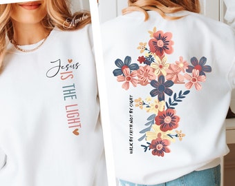 Könnte beinhalten: Weißer Pullover mit einem floralen Kreuzdesign auf der Rückseite und dem Text "Jesus ist das Licht" auf der Vorderseite. Das Kreuz ist von Blumen in den Farben Rosa, Gelb, Blau und Grün umgeben. Der Text ist in einem hellrosa, blauen und grünen Farbschema gehalten.