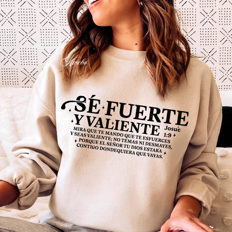 May include: Beige sweatshirt with the text "S&Eacute; FUERTE Y VALIENTE" in black. The text is accompanied by a Bible verse. The sweatshirt has long sleeves and a crew neck. The overall style is casual and comfortable.
