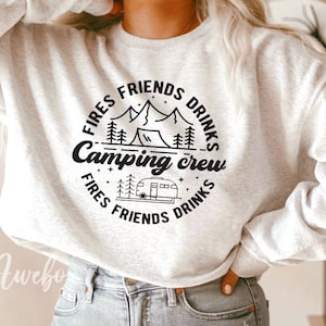 K&ouml;nnte beinhalten: Ein wei&szlig;er Pullover mit einem schwarzen Grafikdesign, auf dem "Fires Friends Drinks Camping Crew Fires Friends Drinks" mit einer Bergszene und einem Wohnmobil steht.