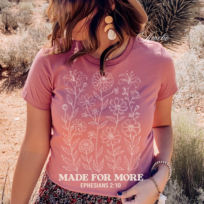 May include: A dusty rose t-shirt featuring a white floral design. The shirt has the text "MADE FOR MORE" and "EPHESIANS 2:10" printed in white. The shirt is short-sleeved and has a crew neck.