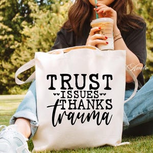 Puede incluir: Bolsa de lona blanca con texto negro que dice "TRUST ISSUES THANKS trauma" con dos peque&ntilde;os corazones a cada lado de la palabra "ISSUES".