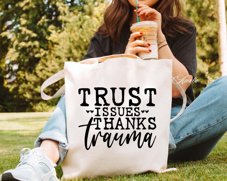 Puede incluir: Bolsa de lona blanca con texto negro que dice "TRUST ISSUES THANKS trauma" con dos peque&ntilde;os corazones a cada lado de la palabra "ISSUES".