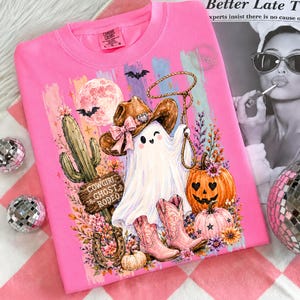 May include: A bright pink t-shirt featuring a graphic of a ghost wearing a brown cowboy hat and pink boots, holding a lasso. The design includes a cactus, a pumpkin, a horseshoe, a full moon, and the text Cowgirl Ghost Rodeo.