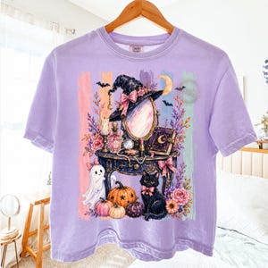 May include: A light purple short-sleeve t-shirt featuring a graphic print of a vanity table with a mirror, a witch hat, a black cat, pumpkins, a ghost, flowers, and a book. The background of the graphic consists of vertical pastel stripes.