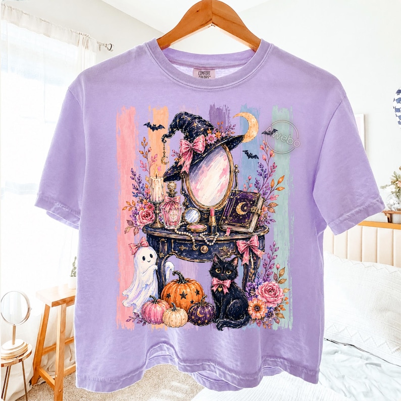 May include: A light purple short-sleeve t-shirt featuring a graphic print of a vanity table with a mirror, a witch hat, a black cat, pumpkins, a ghost, flowers, and a book. The background of the graphic consists of vertical pastel stripes.