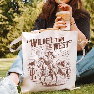 May include: Cream-colored tote bag with the text "Wilder Than The West" and a sepia-toned illustration of a cowgirl riding a horse. The design includes a lasso, desert landscape, and cacti.