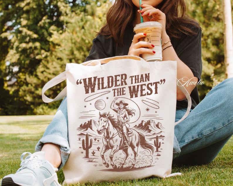 May include: Cream-colored tote bag with the text "Wilder Than The West" and a sepia-toned illustration of a cowgirl riding a horse. The design includes a lasso, desert landscape, and cacti.