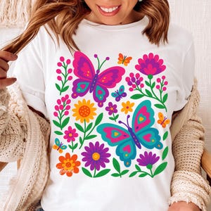 May include: White t-shirt featuring a vibrant floral and butterfly design. The graphic includes pink, purple, teal, orange, and yellow flowers and butterflies. The design is a colorful, eye-catching illustration.