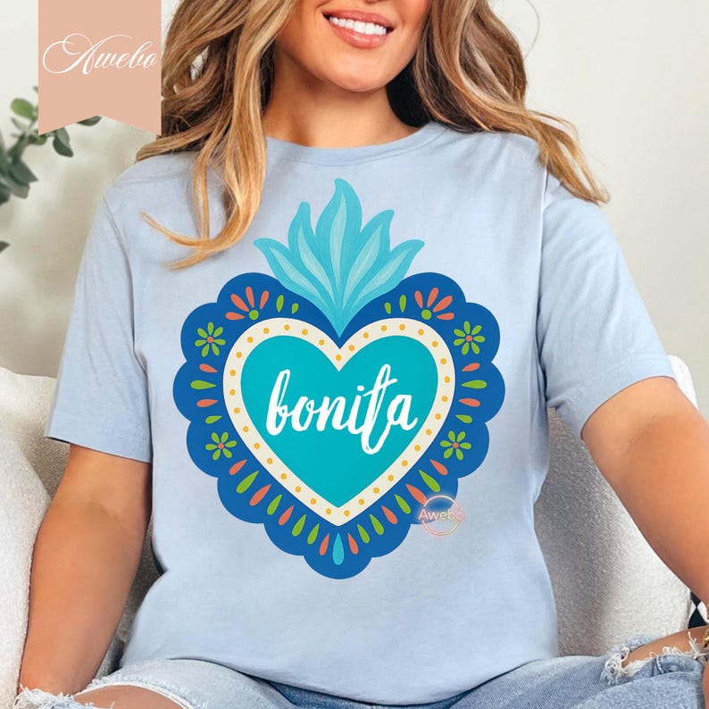 May include: Light blue t-shirt featuring a turquoise heart-shaped design with the word "bonita" in white script. The heart is surrounded by a decorative border with floral and leaf accents in various colors. The shirt has short sleeves.
