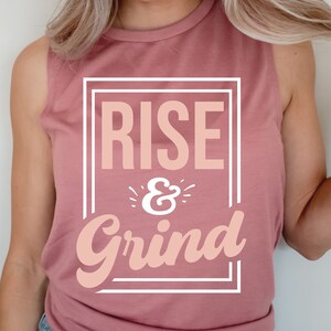 Pode incluir: Uma camiseta rosa sem mangas com o texto "RISE & Grind" em uma caixa branca.