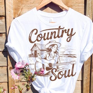 May include: White t-shirt with a brown graphic of a woman playing a guitar, wearing a cowboy hat, and the words "Country Soul". The design includes a barn and a sunset scene. The shirt is hanging on a wooden hanger.