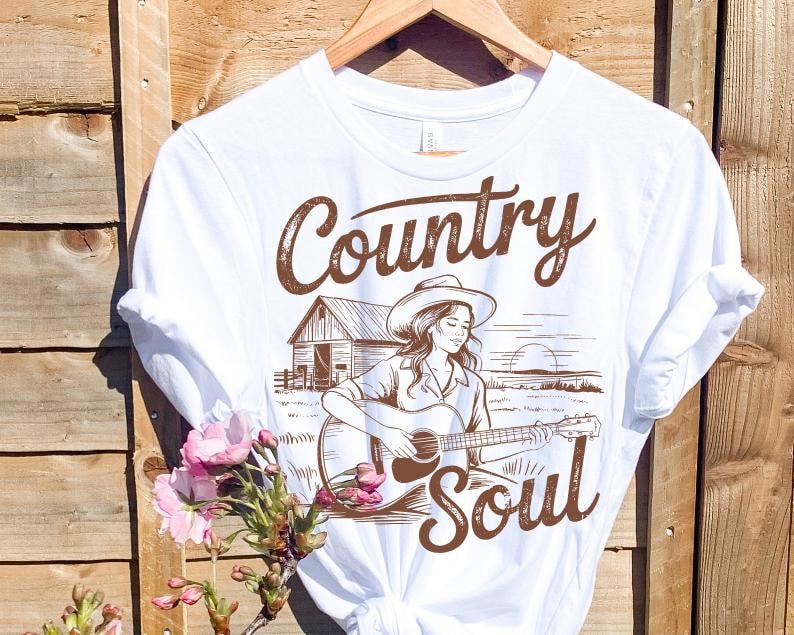 May include: White t-shirt with a brown graphic of a woman playing a guitar, wearing a cowboy hat, and the words "Country Soul". The design includes a barn and a sunset scene. The shirt is hanging on a wooden hanger.