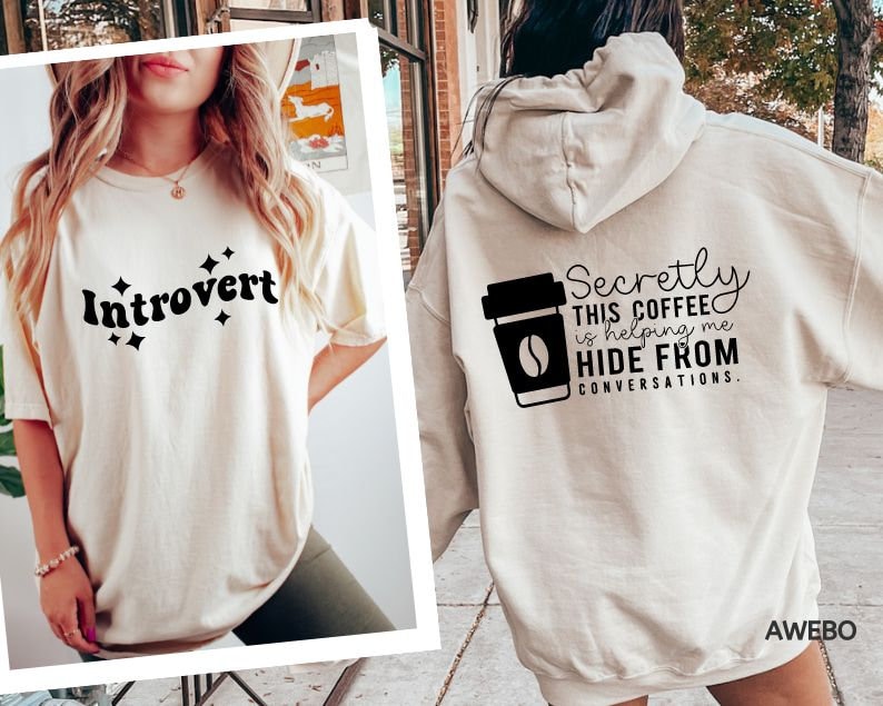 May include: Two cream colored t-shirts with black text. The first shirt says "Introvert" with stars around the word. The second shirt says "Secretly this coffee is helping me hide from conversations."
