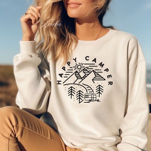 K&ouml;nnte beinhalten: Wei&szlig;er Pullover mit einem schwarzen Grafikdesign aus Bergen, einer Sonne, B&auml;umen und einem Wohnwagen. Der Text "Happy Camper" ist in einem Kreis um das Design geschrieben.