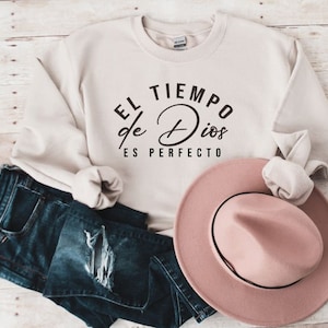 May include: A cream colored sweatshirt with the text "EL TIEMPO de Dios ES PERFECTO" in black lettering. The sweatshirt is folded and displayed with a pink hat and blue jeans.