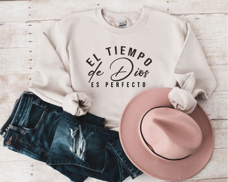 May include: A cream colored sweatshirt with the text "EL TIEMPO de Dios ES PERFECTO" in black lettering. The sweatshirt is folded and displayed with a pink hat and blue jeans.