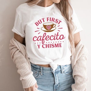 May include: White t-shirt with the text "But first, cafecito y chisme" in pink and brown lettering. The text is surrounded by a stylized coffee cup with a heart design.