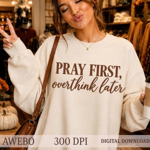 Può includere: Una felpa girocollo color crema con la scritta marrone scuro PRAY FIRST, overthink later stampata sul petto. Il capo presenta maniche lunghe e una vestibilità rilassata, con la tracolla di una borsa marrone che attraversa il davanti.