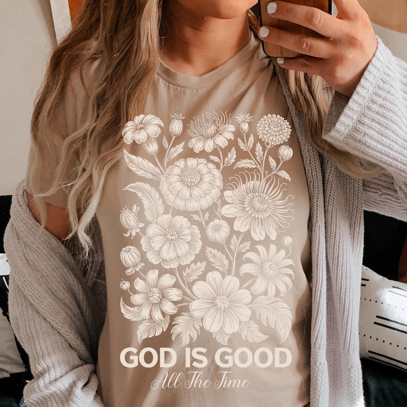 May include: Tan t-shirt featuring a floral design in white. The design includes various flower types and leaves. The text "GOD IS GOOD All The Time" is printed at the bottom of the floral arrangement. The shirt is a neutral color.