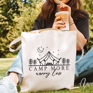 K&ouml;nnte beinhalten: Eine wei&szlig;e Canvas-Tragetasche mit schwarzem Text, der "Camp More Worry Less" lautet. Die Tasche zeigt eine Schwarzwei&szlig;illustration eines Zeltes, Sterne und B&auml;ume.